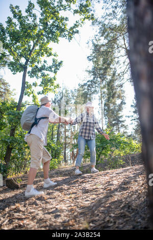 Husband helping wife. Supporting husband wearing backpack helping wife hiking in forest Stock Photo