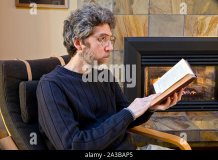 Man reading a book by the fire at home. Middle aged Caucasian male reads a book at home. Stock Photo