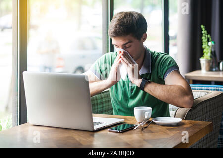 Young cold sick businessman in green t-shirt sitting at work, sneezing and cleaning his nose with tissue napkin. business and helthcare concept. indoo Stock Photo