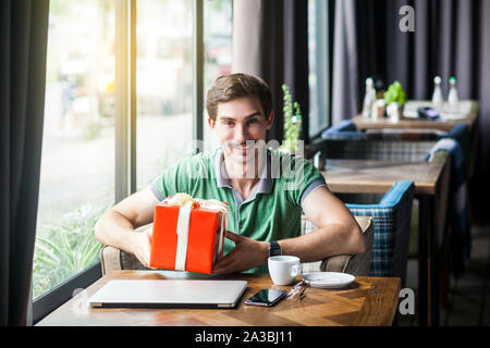 Young happy businessman in green t-shirt sitting and holding big red gift box and looking at camera with toothy smile. business and freelancing concep Stock Photo