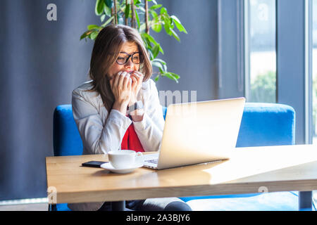 Portrait of nervous beautiful stylish brunette young woman in glasses sitting and looking at laptop monitor, bitting her nails and worry or panic. ind Stock Photo