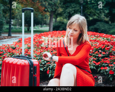 blonde girl dressed in red, waiting sitting on a staircase and next to a travel suitcase Stock Photo