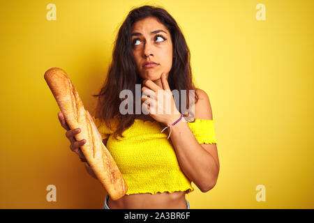 Young beautiful baker woman holding bread standing over isolated yellow background serious face thinking about question, very confused idea Stock Photo