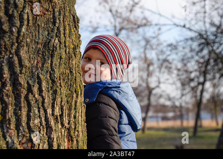 A little boy is hiding behind a big tree. A child peeks out from behind a tree trunk. Stock Photo