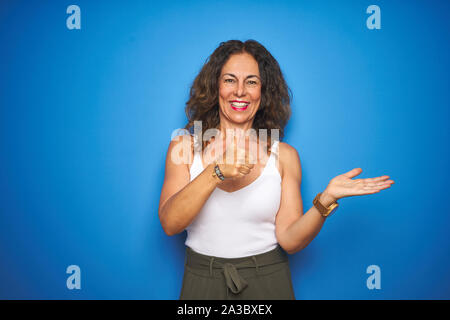 Middle age senior woman with curly hair standing over blue isolated background Showing palm hand and doing ok gesture with thumbs up, smiling happy an Stock Photo