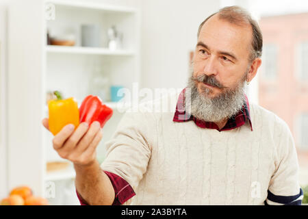 Portrait of bearded senior man holding fresh bell peppers while cooking vegetables in kitchen, copy space Stock Photo