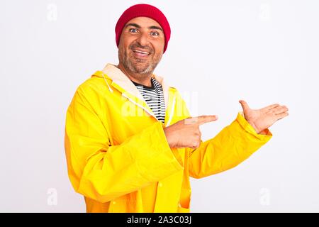 Middle age man wearing rain coat and woolen hat standing over isolated white background amazed and smiling to the camera while presenting with hand an Stock Photo