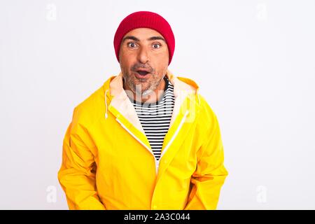 Middle age man wearing rain coat and woolen hat standing over isolated white background afraid and shocked with surprise expression, fear and excited Stock Photo