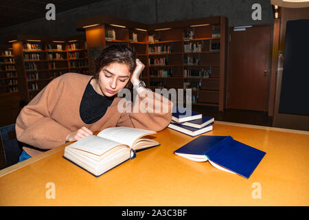 Young brunette thoughtfully reads book in library Stock Photo