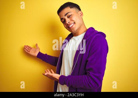 Young brazilian man wearing purple sweatshirt standing over isolated yellow background Inviting to enter smiling natural with open hand Stock Photo