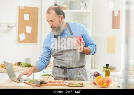 Portrait of bearded senior man using laptop while cooking dinner in cozy kitchen at home, copy space Stock Photo