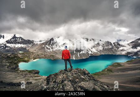 Man in red jacket is looking at Ala-Kul Lake in the Tien Shan mountains with white foggy clouds in Karakol national park, Kyrgyzstan