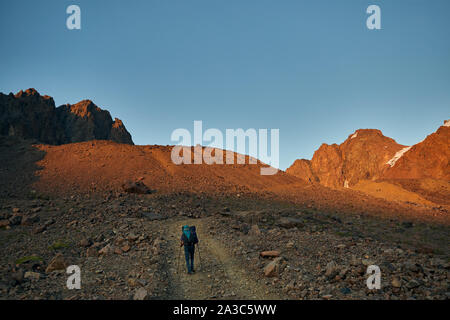 Hiker with big backpack is walking on the road with spectacular mountains at sunset Stock Photo