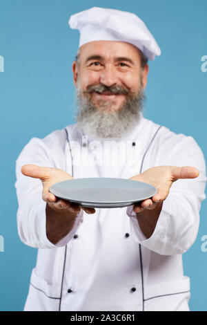 Portrait of bearded senior chef holding empty plate and smiling at camera while standing against blue background, mock up template, focus on foreground Stock Photo