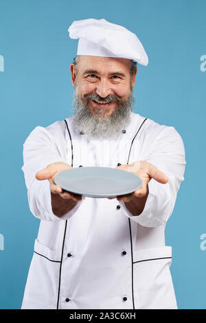 Portrait of bearded senior chef holding empty plate and smiling at camera while standing against blue background, mock up template Stock Photo