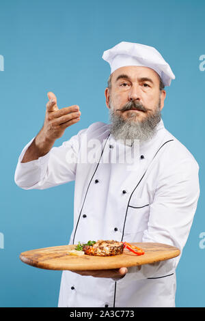Waist up portrait of charismatic senior chef holding beautiful dish on wooden platter while standing against blue background and looking at camera Stock Photo