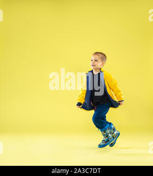 A full length portrait of a bright fashionable boy in a raincoat running and having fun on yellow studio background. Autumn and spring fashion for kids. Cute stylish boy in colorful clothes. Stock Photo