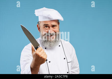 Portrait of crazy-looking bearded chef holding sharp knife and smiling at camera while standing against blue background, copy space Stock Photo