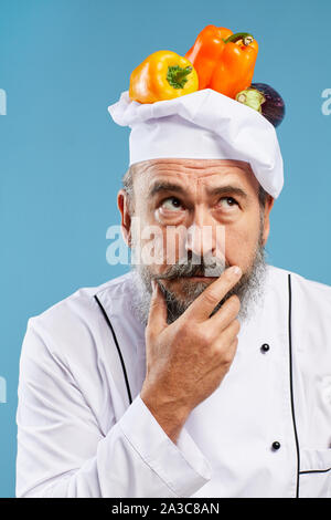 Portrait of charismatic bearded chef looking away pensively while posing against blue background with fresh vegetables on top of hat Stock Photo