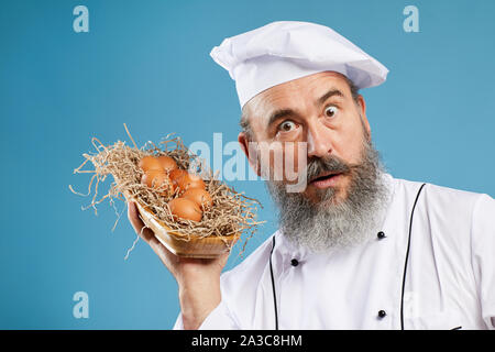 Portrait of charismatic bearded chef holding basket with fresh eggs while posing against blue background and looking shocked, copy space