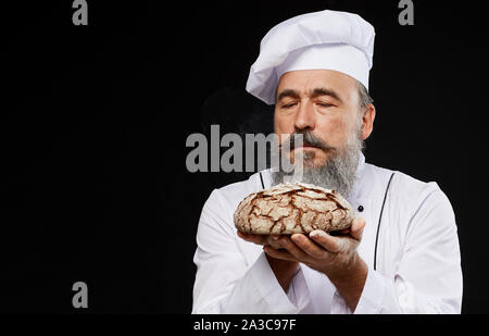 Portrait of charismatic bearded baker holding fresh bread loaf lovingly while standing against black background, copy space Stock Photo