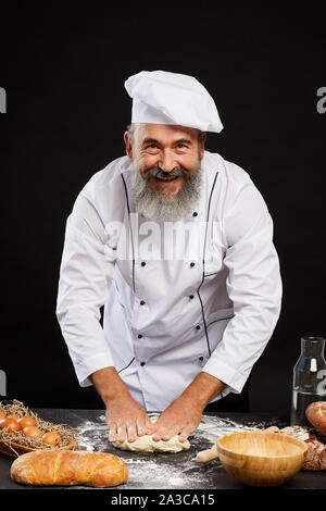 Portrait of charismatic bearded baker making fresh bred and smiling cheerfully at camera while standing against black background Stock Photo