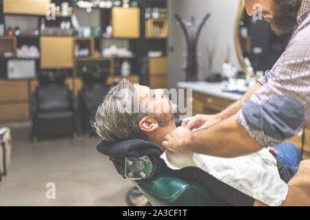 Hairdresser preparing client for shaving with towel. Man visiting barber shop. Stock Photo