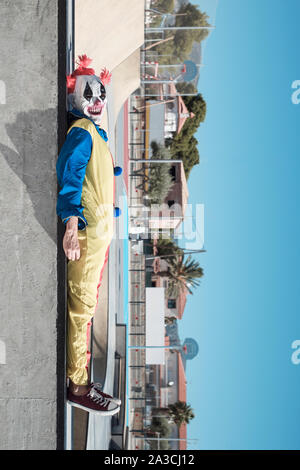 a scary clown, wearing a colorful yellow, red and blue costume, lying down in an outdoor public sports complex Stock Photo