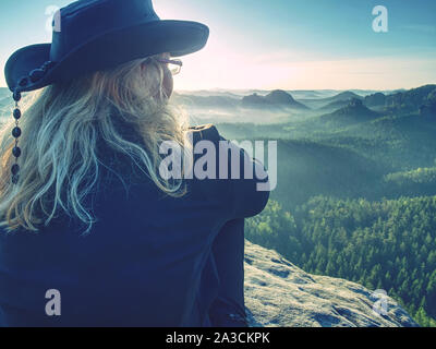 Young blond hair girl hiker sitting on a cliff and enjoy the sunset foggy rocky mountains in Saxon Switzerland, Germany. Stock Photo