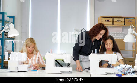 Cutting and sewing courses. Seamstress teacher teaches her students to sew on sewing machines in workshop. Dressmaker talking with woman explaining and showing them how to sew clothes and make seams. Stock Photo