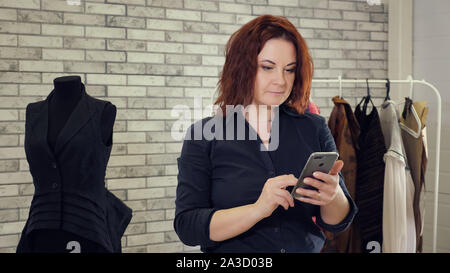 Stylist is working in sewing factory using mobile phone and typing message. Mature woman fashion designer with mobile phone types message in tailoring studio among clothes and mannequins. Stock Photo