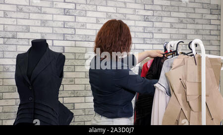Working in tailoring business factory. Unrecognizable woman fashion designer in workshop chooses clothes hanging on hangers in atelier. She evaluates ready-to-run clothing models in production. Stock Photo