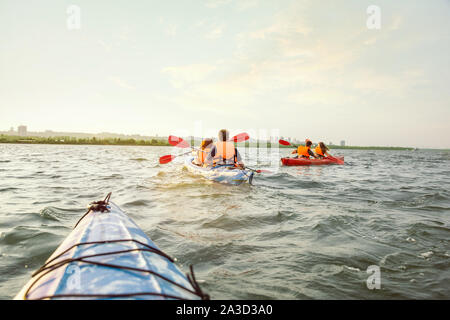 Happy young caucasian group of friends kayaking on river with sunset in the backgrounds. Having fun in leisure activity. Happy male and female model laughting on the kayak. Sport, relations concept. Stock Photo
