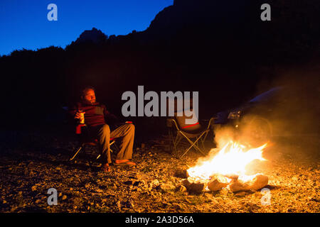 Sitting at a glowing camp fire with a beer in wilderness at night, freedom camping in beautiful nature, Valbona Valley, Albania Stock Photo