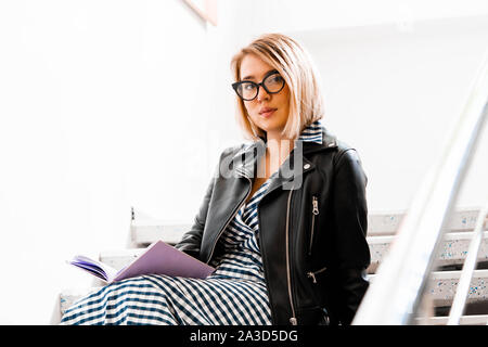 Young attractive blond woman makes some notes in violet notepad at library and sitting on ladder steps. Stock Photo