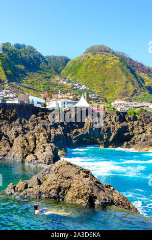 Lady swimming in natural swimming pools in the Atlantic ocean, Madeira Island, Portugal. Made up of volcanic rock, into which sea flows naturally. City Porto Moniz on the mountain in the background. Stock Photo