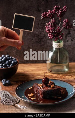 Caucasian male hand holding small blank chalkboard sign over homemade vegan chocolate brownie with chocolate sauce, served on blue plate. Copy space Stock Photo