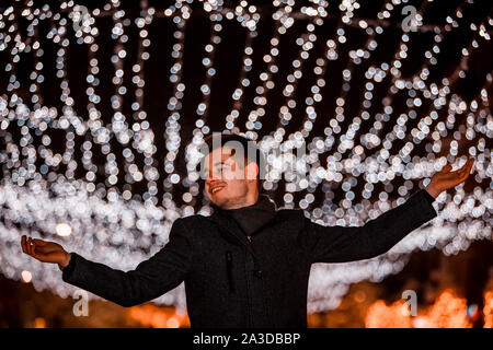 Free young man with outstretched arms looking up while being excited about winter weather at decorated city street at night Stock Photo