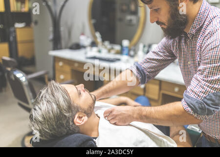 Barber covering client with towel before giving him a shave at barbershop. Stock Photo