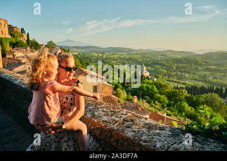From above cute girl pointing at picturesque Tuscany landscape while young pretty mother in sunglasses standing and leaning on parapet carrying daught Stock Photo