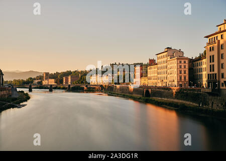 Old buildings located on shore of calm river and bridge against cloudless sundown sky in Tuscany, Italy Stock Photo