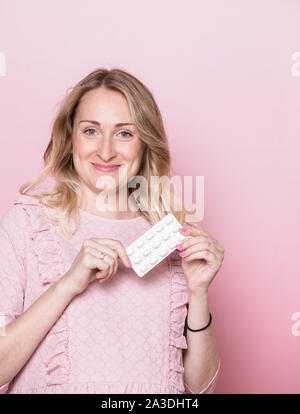 Young pregnant woman wearing pink dress holding blister pack of medicine pills looking at camera smiling. Studio shot on pink background. Prenatal med Stock Photo