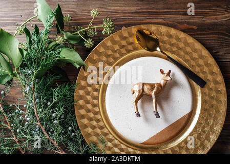 Holiday place setting and toy golden reindeer decoration on the plate surrounded by natural branches, on wooden background. Stock Photo