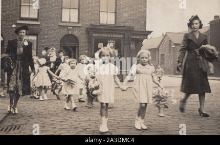 Vintage Early 20th Century Photograph Showing Pretty Young Girls Holding Floral Baskets Being Led Across a Cobbled Street By Ladies. Grays Quality Repairs Shop In The Background. Stock Photo