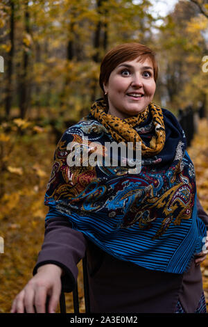 Fat girl on the bridge surprised look around maple leaves and autumn forest at sunset in a painted shawl Stock Photo