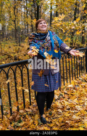 Fat girl on the bridge rejoices redhead around maple leaves fly and autumn forest at sunset in a painted shawl Stock Photo