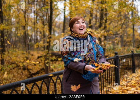 Fat girl on the bridge rejoices around flying maple leaves and autumn forest at sunset in a painted shawl Stock Photo