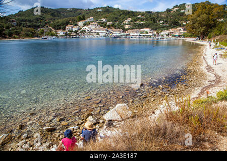 Two people sitting on the sea shore towards the Greek fishing village of  Agios Sstefanos on the Greek Island of Corfu Greece Stock Photo