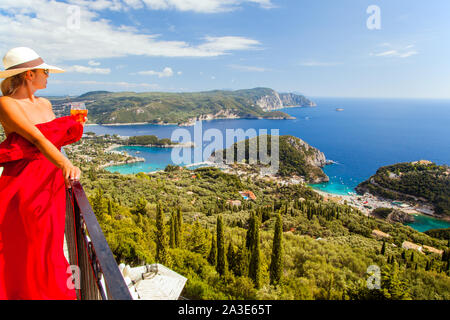 Woman in long red dress  wearing a hat holding a cocktail standing on a balcony at Bella Vista above the Greek resort of Paleokastritsa Corfu Greece Stock Photo