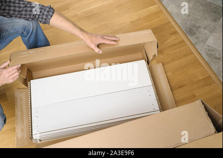 A man is unpacking a cardboard box with furniture. On the floor there is a box with white boards. Stock Photo